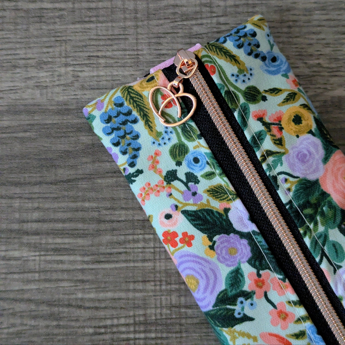 A close-up of a floral zippered pouch with a mint green background and a multicolor print of flowers and leaves, featuring a rose gold zipper pull with a heart and interlocking rings design, set against a gray wood-textured background.