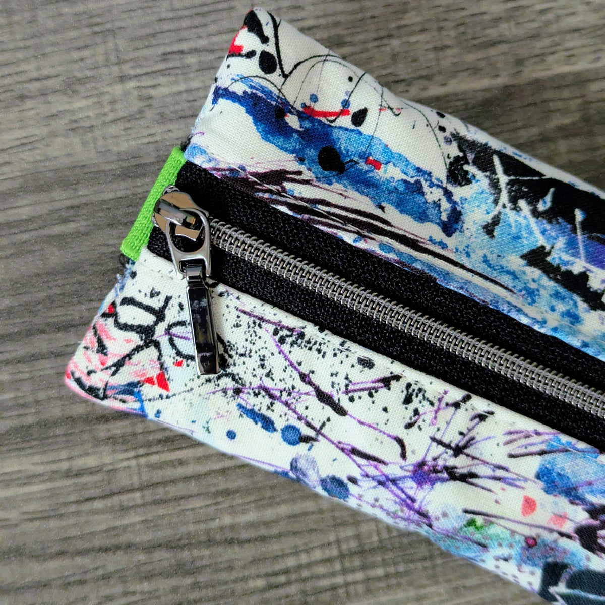 A close-up of a zippered pouch with a colorful abstract paint splatter pattern on a white background, resting on a gray wood-textured surface.