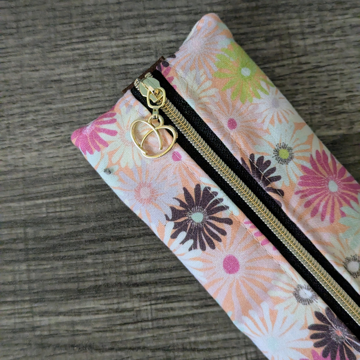 A close-up of a zippered fabric pouch with a colorful floral print, featuring pink, green, and purple flowers on a white background, with a gold zipper and a gold heart-shaped zipper pull, set against a dark gray wood-grain background.