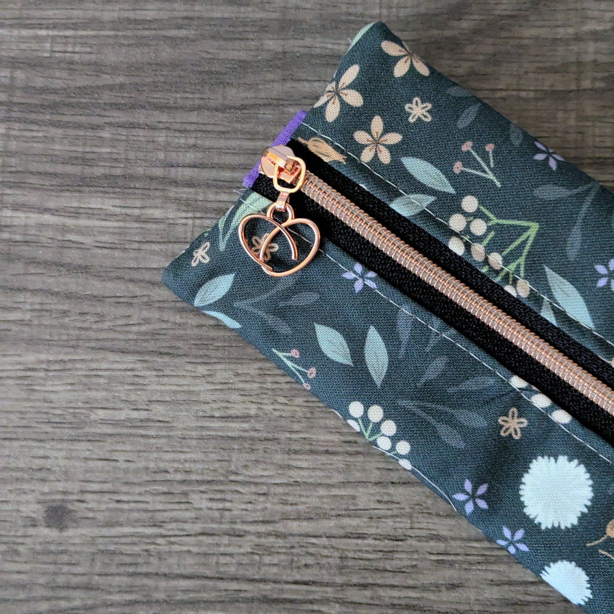 A close-up of a fabric zipper pouch with a dark teal floral pattern, featuring a rose gold zipper and a matching rose gold heart-shaped zipper pull, shown on a gray wood-textured background.
