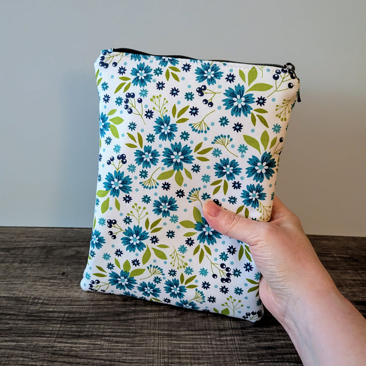 Floral-patterned pouch held by a hand on a wooden surface with a neutral background
