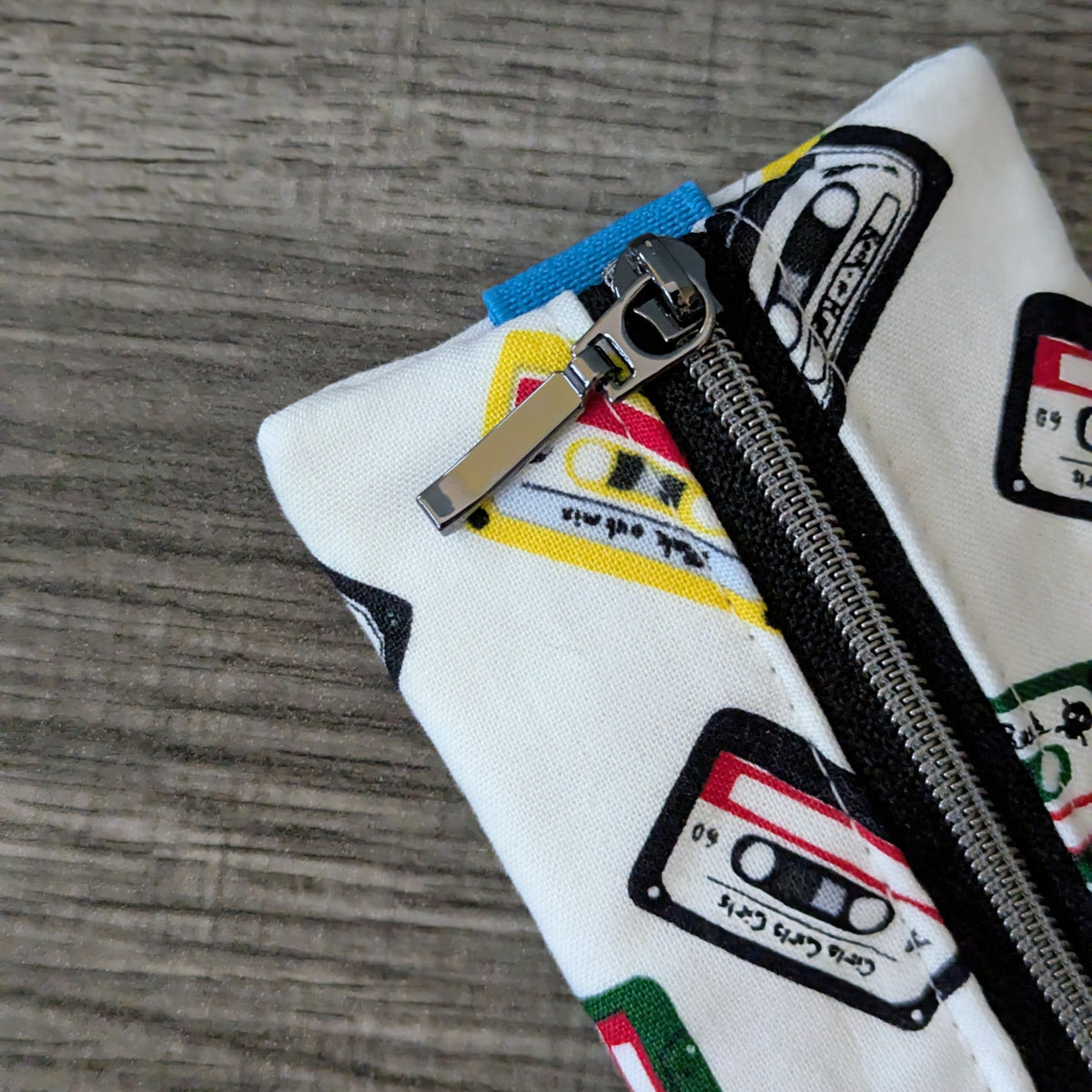A close-up of a white fabric zippered pouch decorated with retro cassette tape graphics, resting on a gray wood-grain surface.