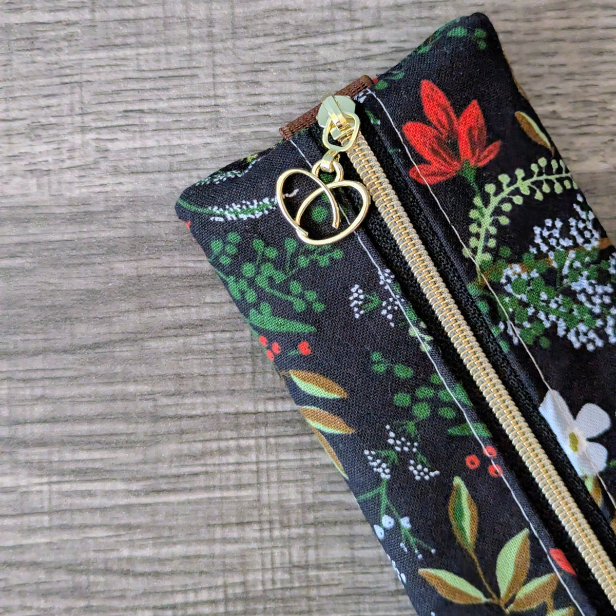 A close-up of a zipper pouch with a black background and a colorful floral print, featuring a gold heart-shaped zipper pull, set against a gray wood-textured background.