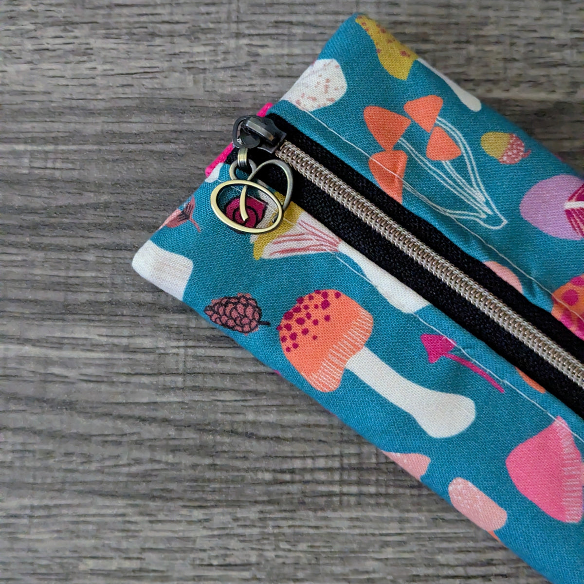 A close-up of a fabric zipper pouch with a colorful mushroom print on a teal background, featuring a black zipper and a peace-sign charm, set on a gray wood-textured surface.