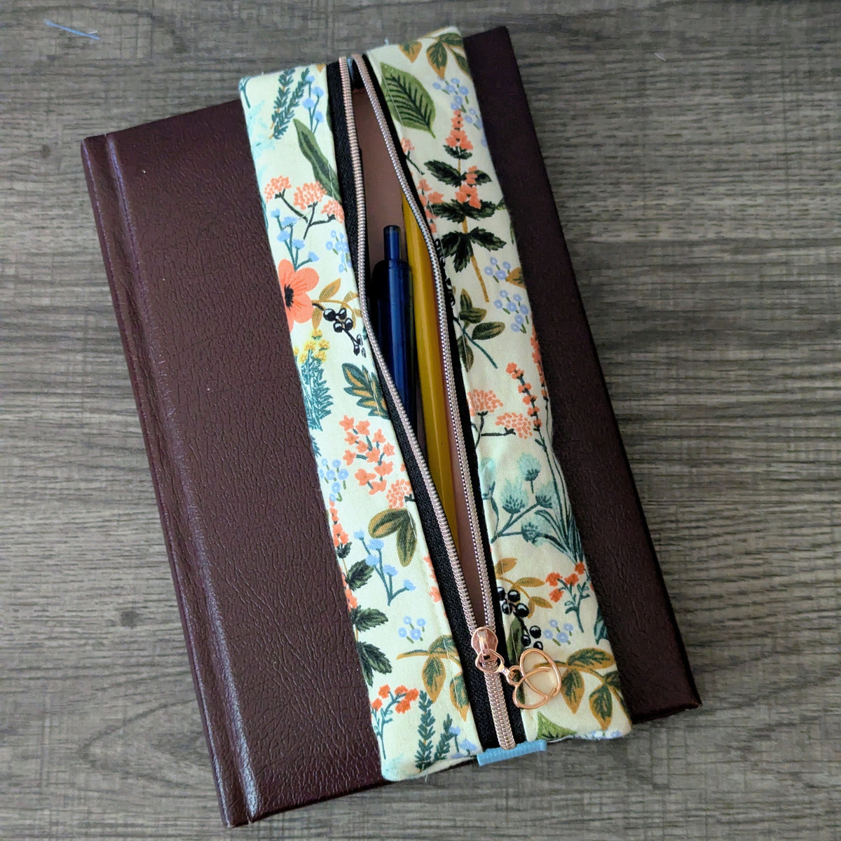 A floral zippered pouch with a brown faux-leather side panel, shown open on a gray wood-textured surface with pens visible inside.