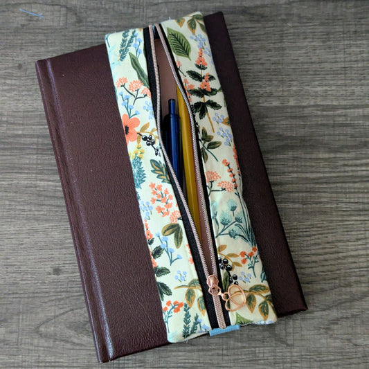 A floral zippered pouch with a brown faux-leather side panel, shown open on a gray wood-textured surface with pens visible inside.