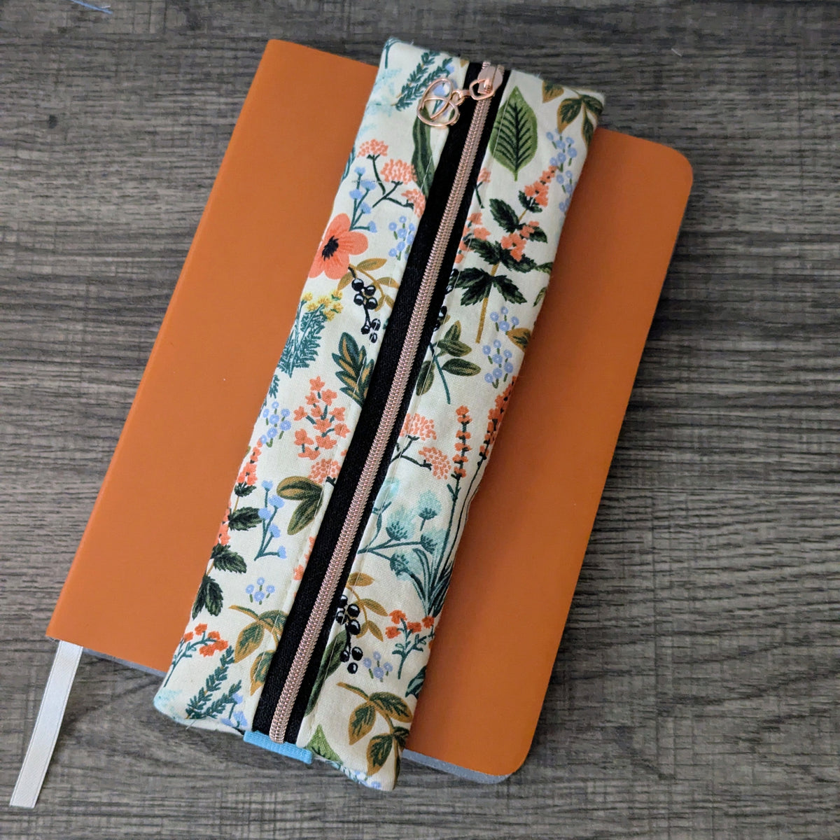 A floral fabric zippered pouch with a black center stripe and rose gold zipper pulls, laid diagonally over an orange notebook on a gray wood-textured surface.