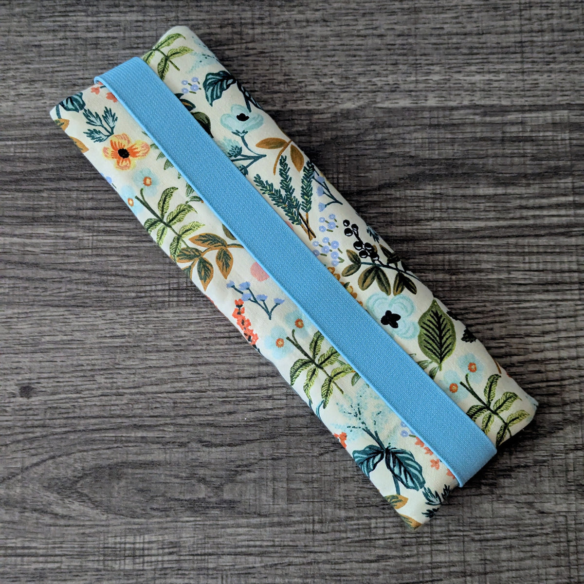 A fabric-covered pencil case with a light blue elastic band, featuring a botanical floral print in green, orange, and blue on a cream background, shown on a dark wood surface.