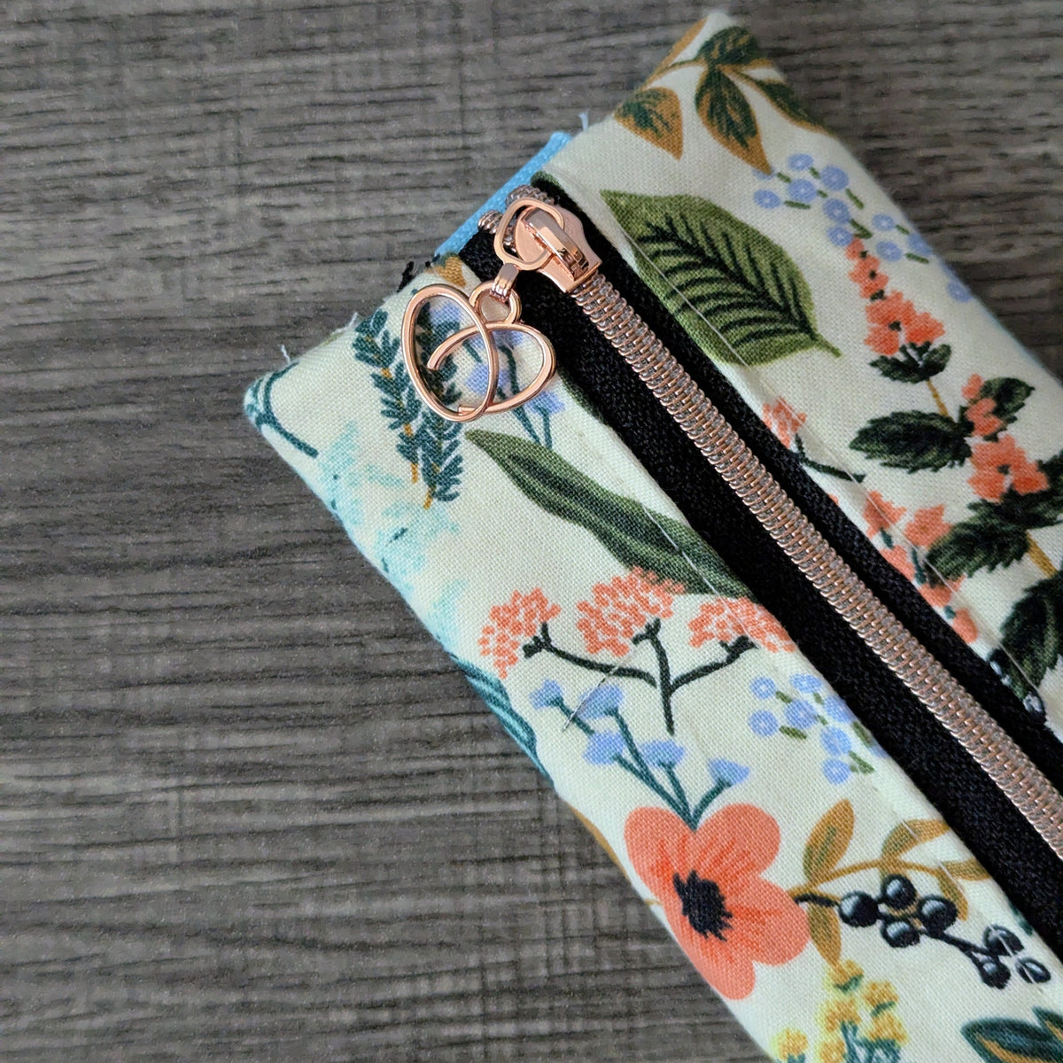 A close-up of a fabric zipper pouch with a floral print, featuring a cream background with green leaves and orange, blue, and yellow flowers. The pouch has a rose gold heart-shaped zipper pull and a black zipper track, shown on a gray wood-textured background.