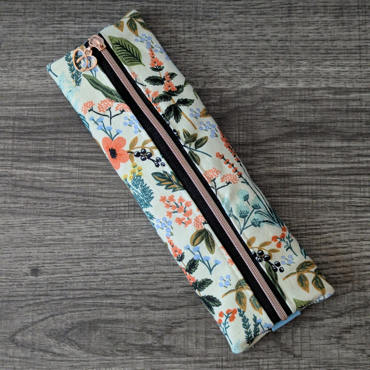 A long, narrow zippered pouch with a colorful floral print on a light cream background, featuring a black zipper with a rose gold metal pull, placed diagonally on a gray wood-textured surface.