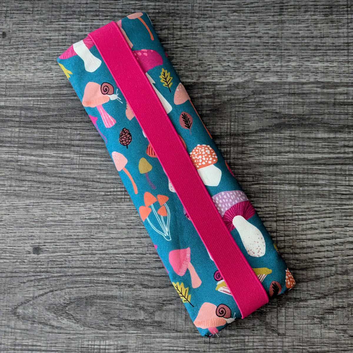 A rectangular fabric pouch with a colorful mushroom print on a teal background and a bright pink elastic band, photographed on a gray wood surface.