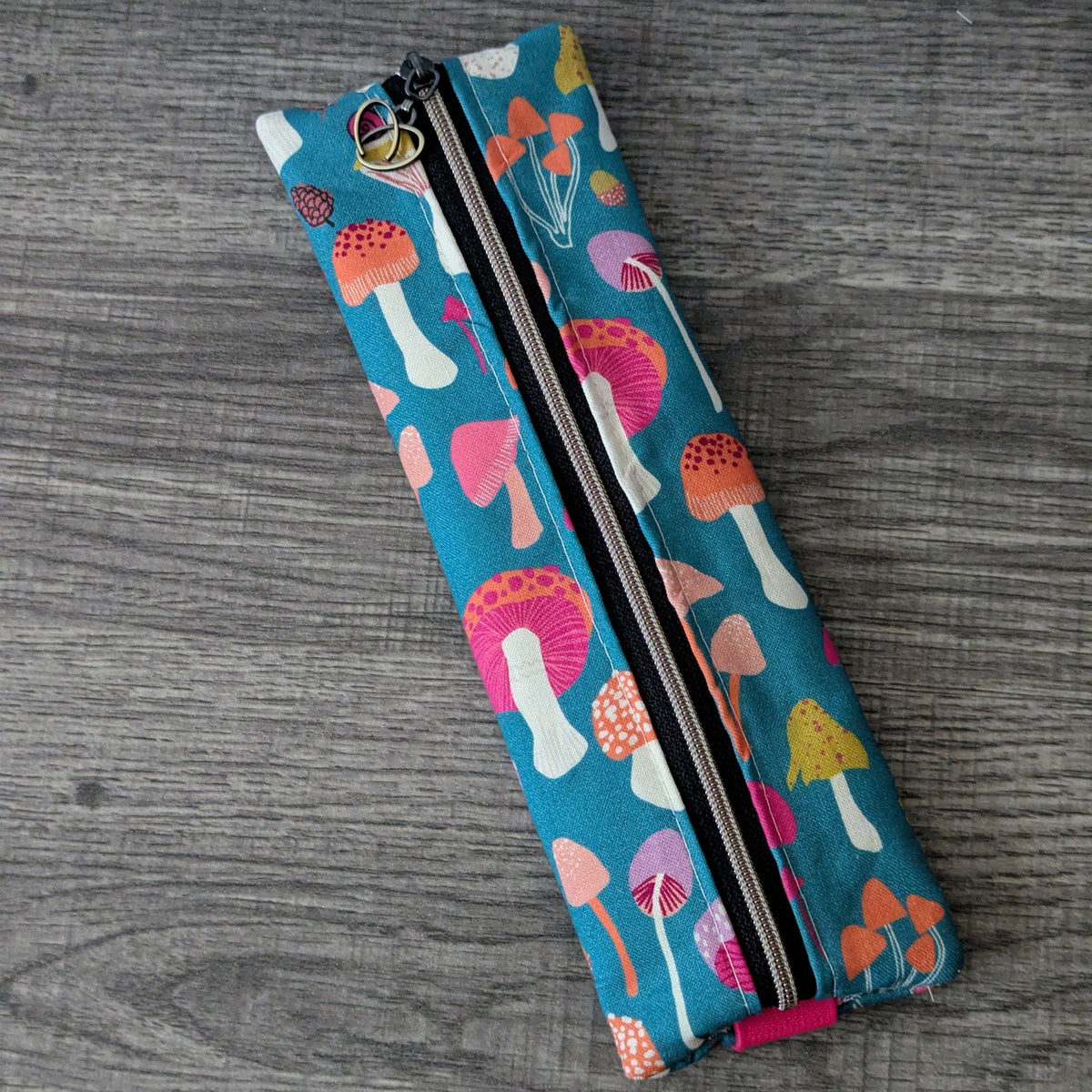 A slim zippered fabric pouch with a colorful mushroom print on a teal background, placed diagonally on a gray wood-textured surface.