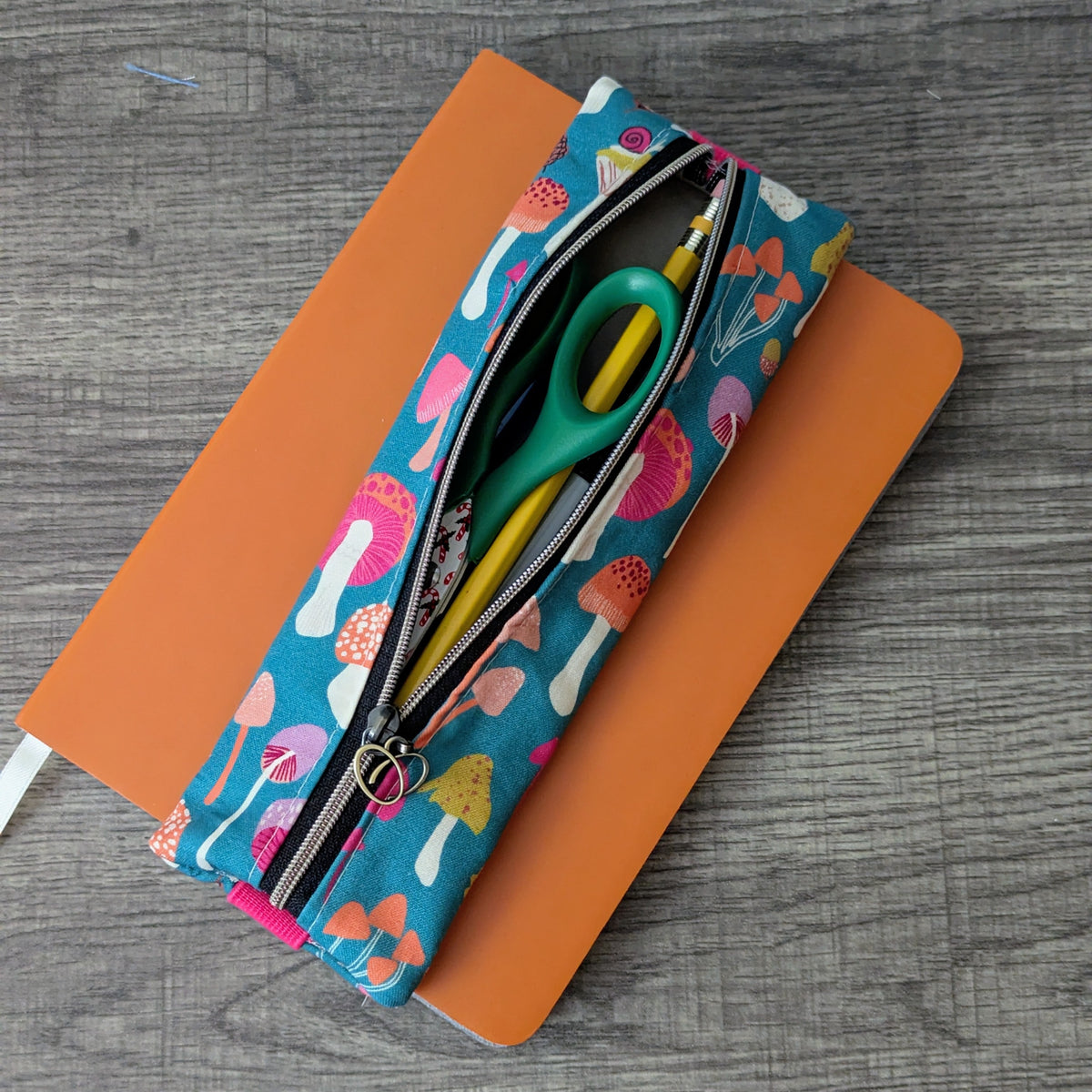 A fabric pencil case with a colorful mushroom print lies open on top of an orange notebook, with green-handled scissors and yellow pens visible inside, set on a gray wood-textured surface.