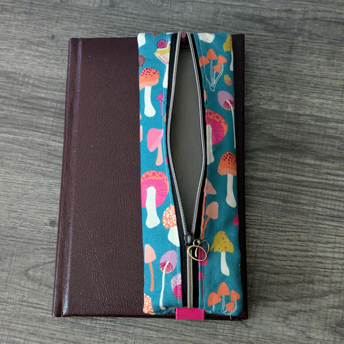 A zippered notebook cover or journal sleeve lying flat on a gray wood-grain surface. The left side is dark brown faux leather, and the center features a vertical panel with a colorful mushroom print on teal fabric and a black zipper running through it.