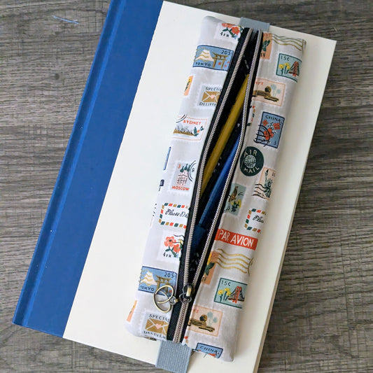 A zippered pouch with a colorful vintage postage stamp pattern, resting on top of a white and blue notebook on a gray wood-textured surface.