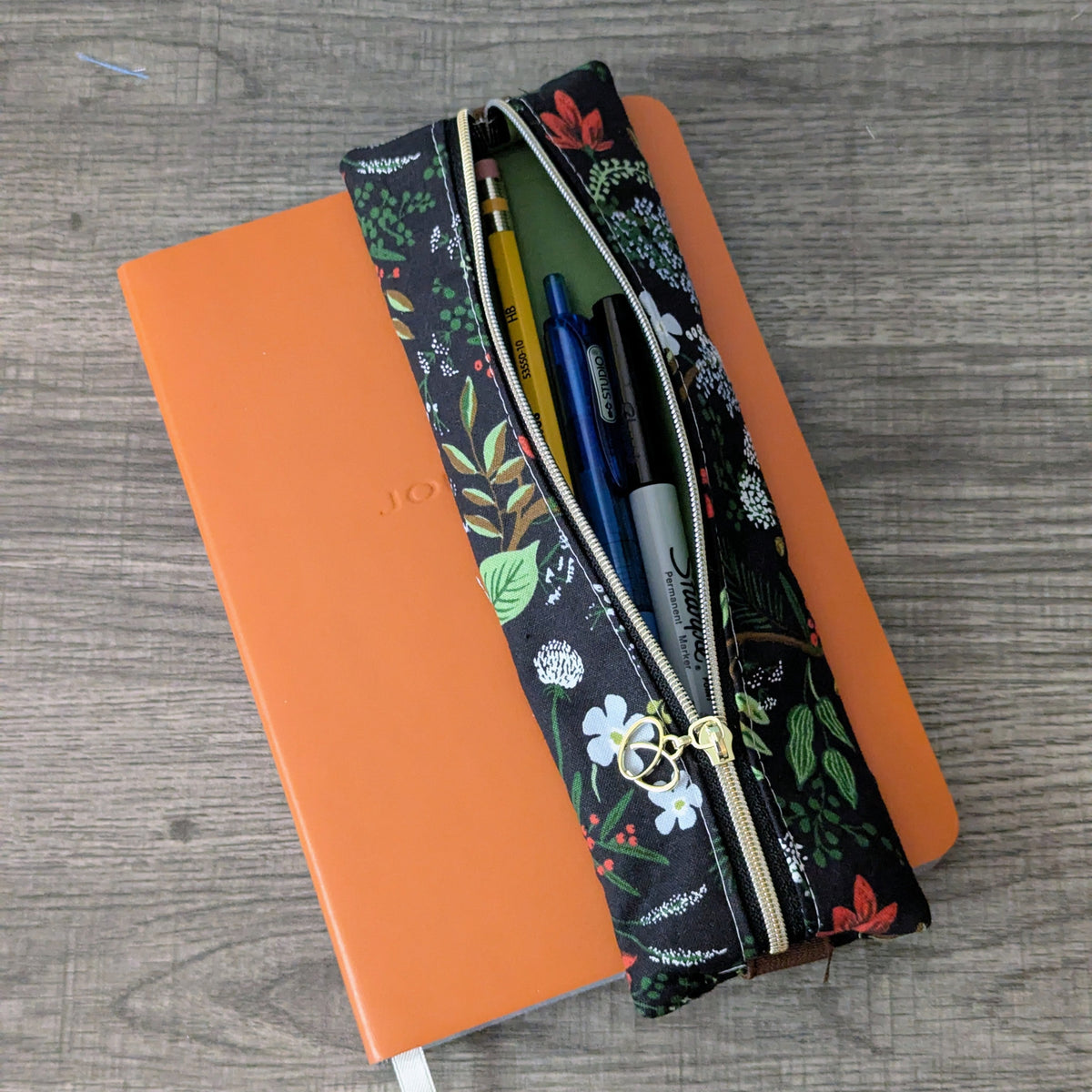 A floral-patterned zippered pencil case with a gold zipper, shown open and filled with pens, resting on top of an orange notebook on a gray wood-textured surface.