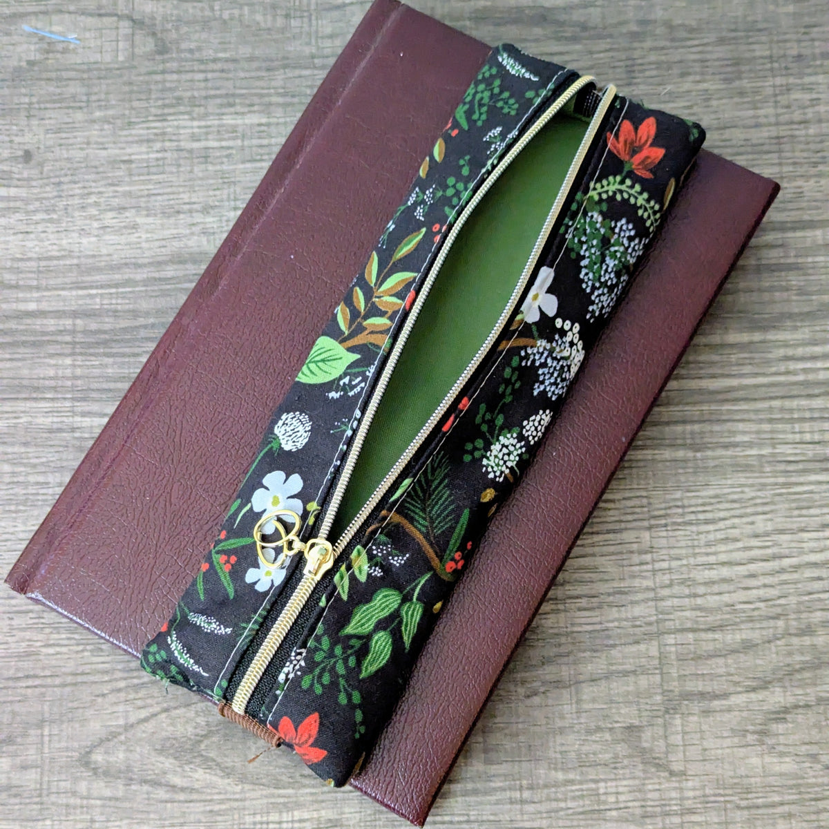 A floral-patterned zipper pouch with a gold zipper, shown open and resting on top of a dark brown book on a gray wood-textured surface.