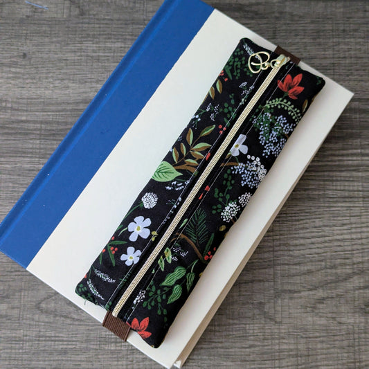 A floral-patterned zipper pouch with a black background and multicolor botanical print, resting diagonally on top of a white book with a blue spine on a gray wood-textured surface.
