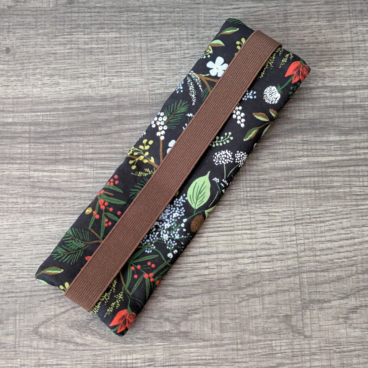 A rectangular fabric pouch or case with a brown elastic band, featuring a black background with a colorful floral and botanical print, placed diagonally on a gray wood-textured surface.