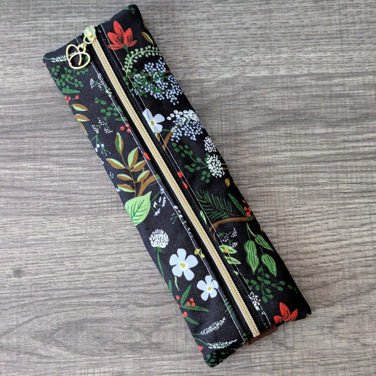 A long, narrow fabric zipper pouch with a black background and a colorful botanical print, featuring green leaves, white and red flowers, and a gold zipper.