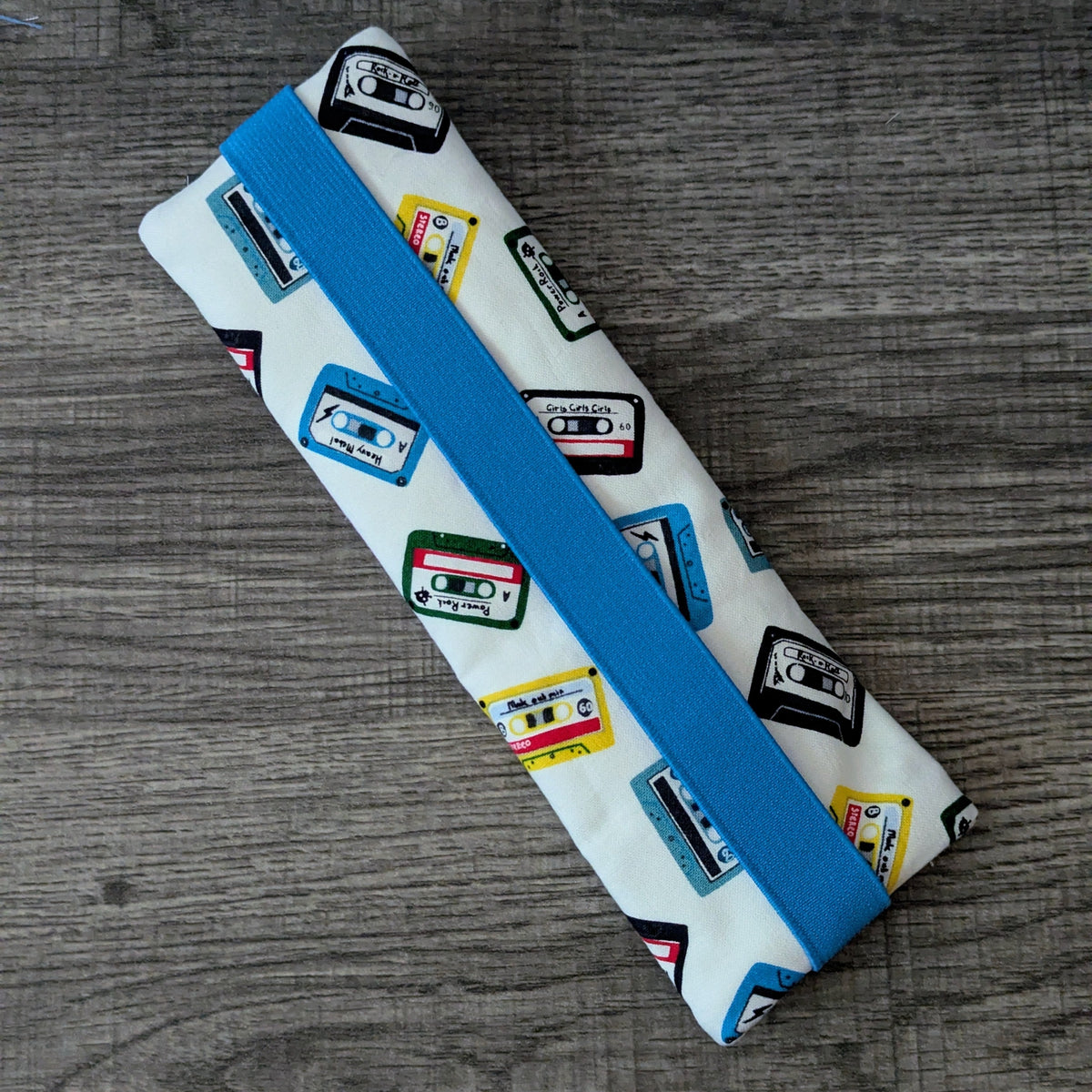 A rectangular fabric pouch or case with a white background printed with colorful cassette tape motifs and a bright blue elastic band, photographed on a gray wood surface.