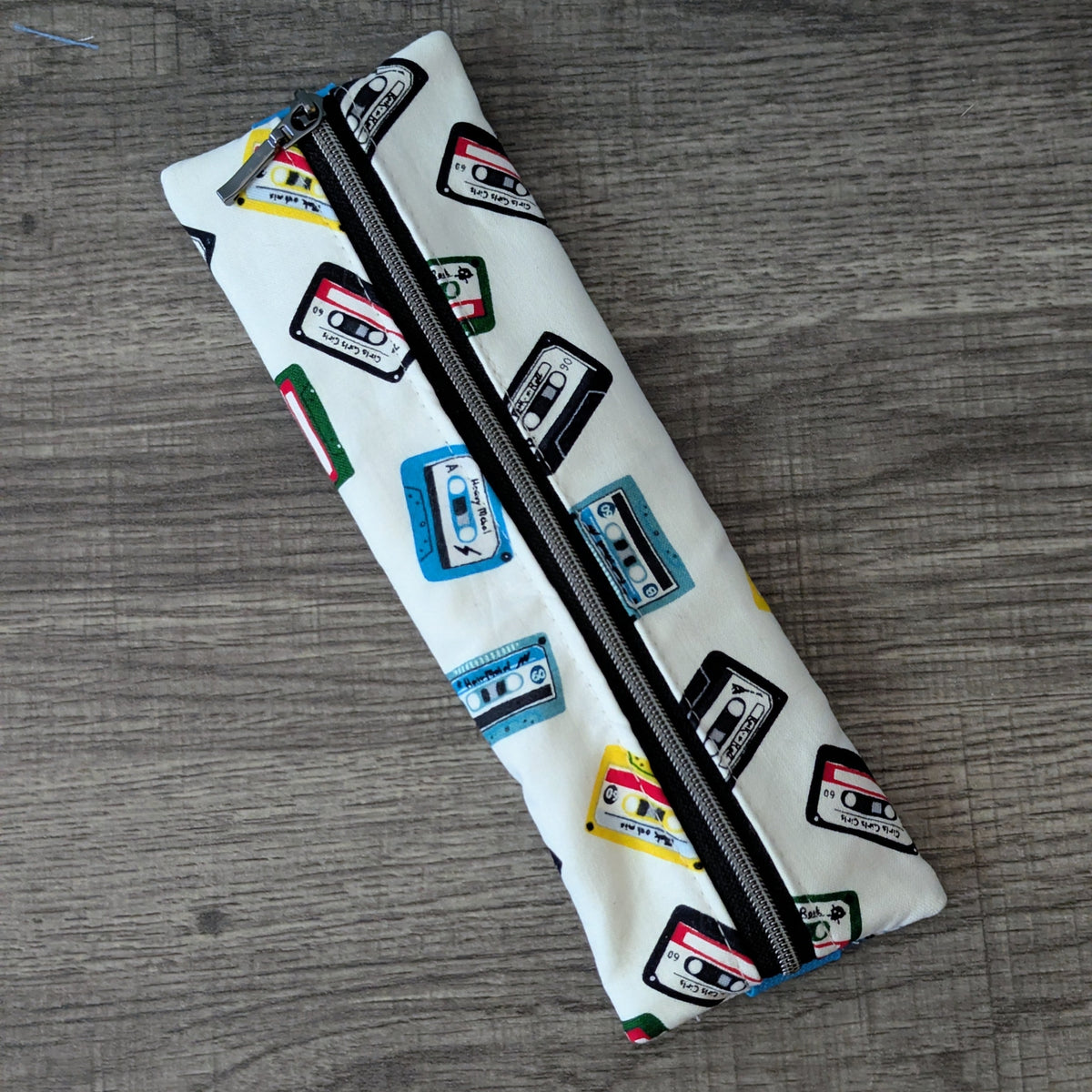 A white zippered pencil case featuring a colorful pattern of retro cassette tapes, placed diagonally on a gray wood-textured surface.