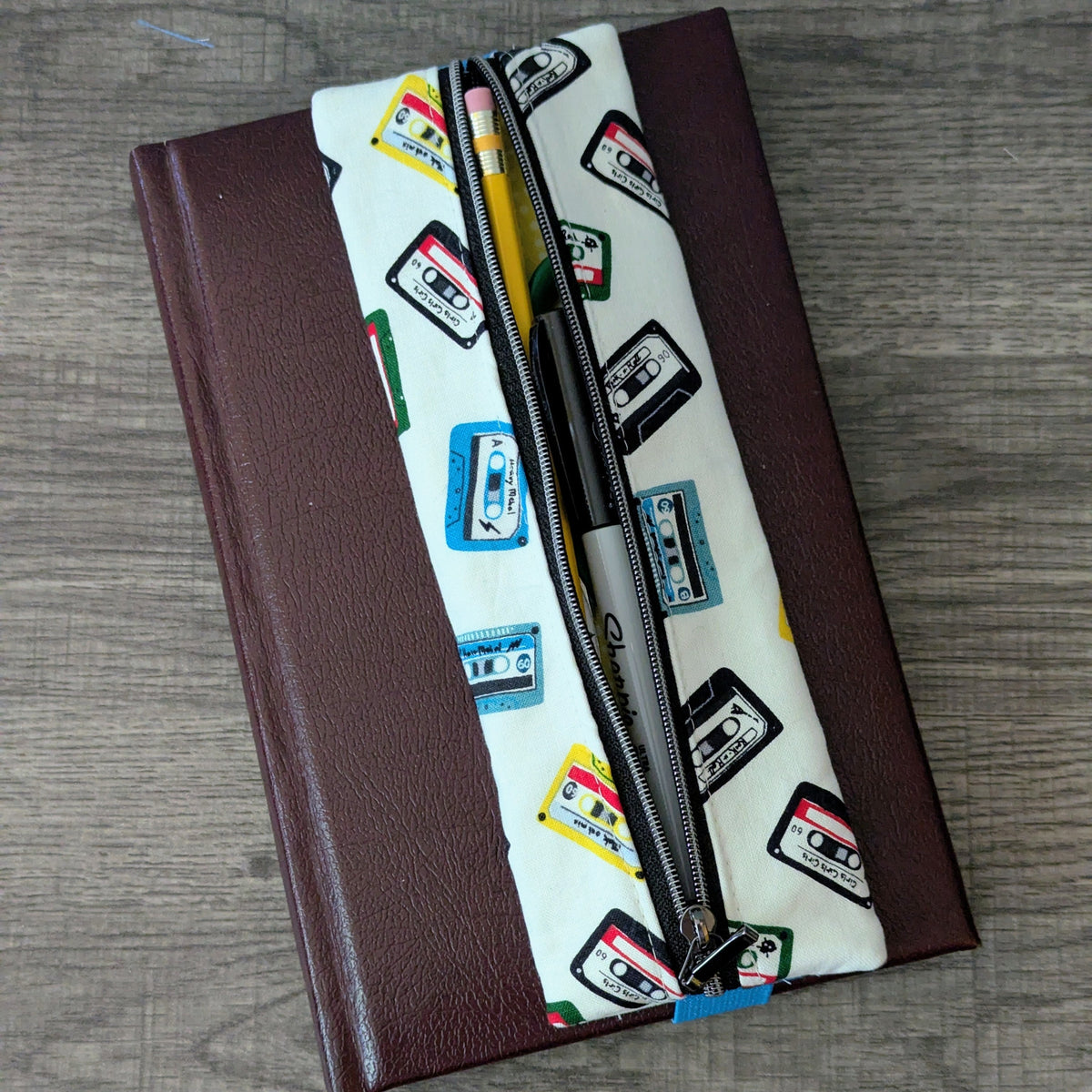 A zippered pouch with a white background and colorful cassette tape pattern, placed diagonally over a dark brown hardcover notebook on a gray wood-textured surface.
