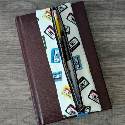 A zippered pouch with a white background and colorful cassette tape pattern, placed diagonally over a dark brown hardcover notebook on a gray wood-textured surface.