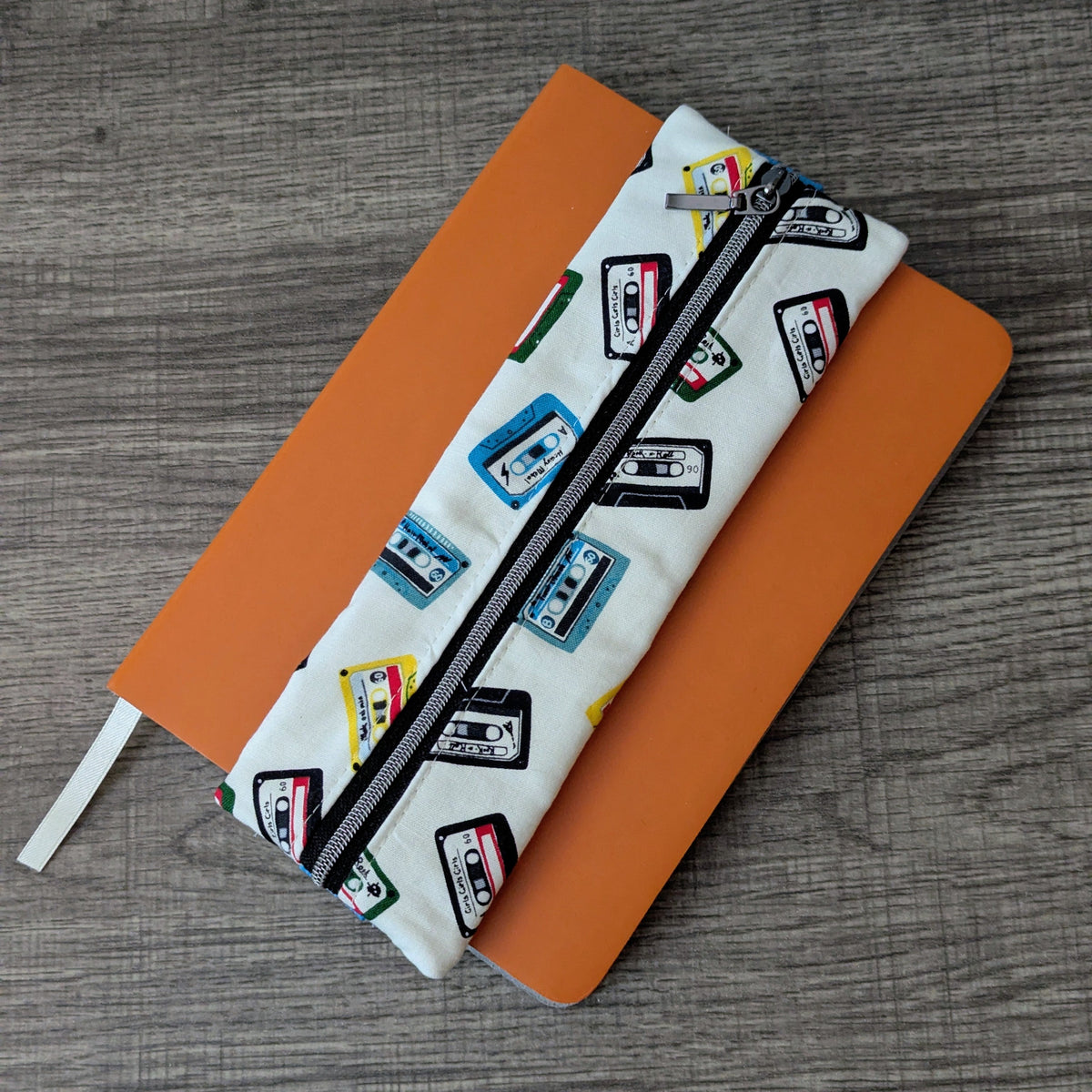 A white zipper pouch with a colorful cassette tape pattern, placed diagonally over an orange notebook on a gray wood-grain tabletop.