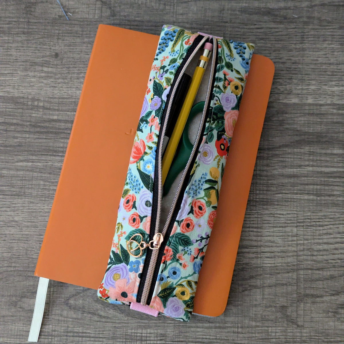 A floral zippered pencil case holding a yellow pencil, resting on top of an orange notebook over a gray wood-grain surface.
