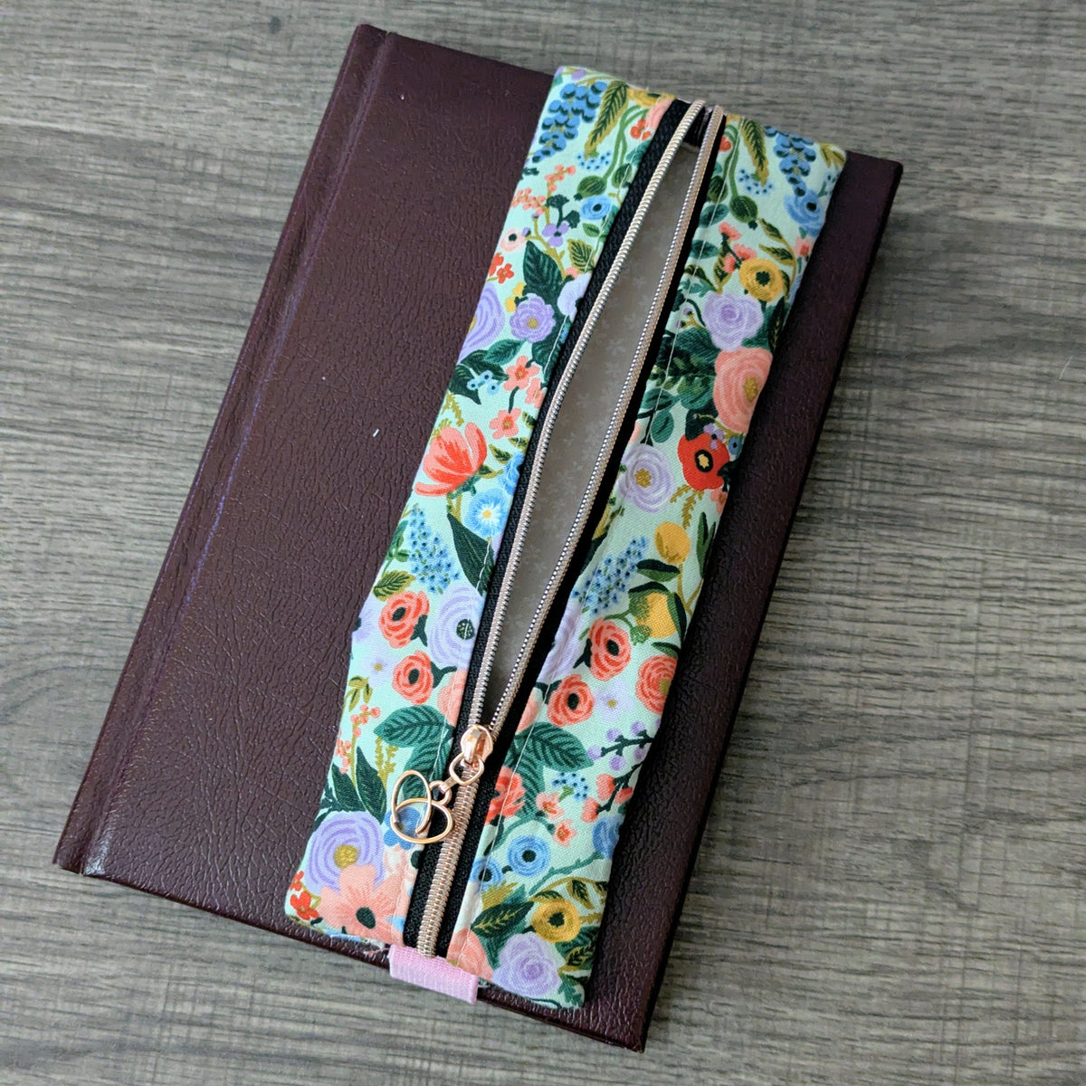 A dark brown leather-bound notebook or journal with a colorful floral-patterned fabric pencil case, placed on a gray wood-textured surface.