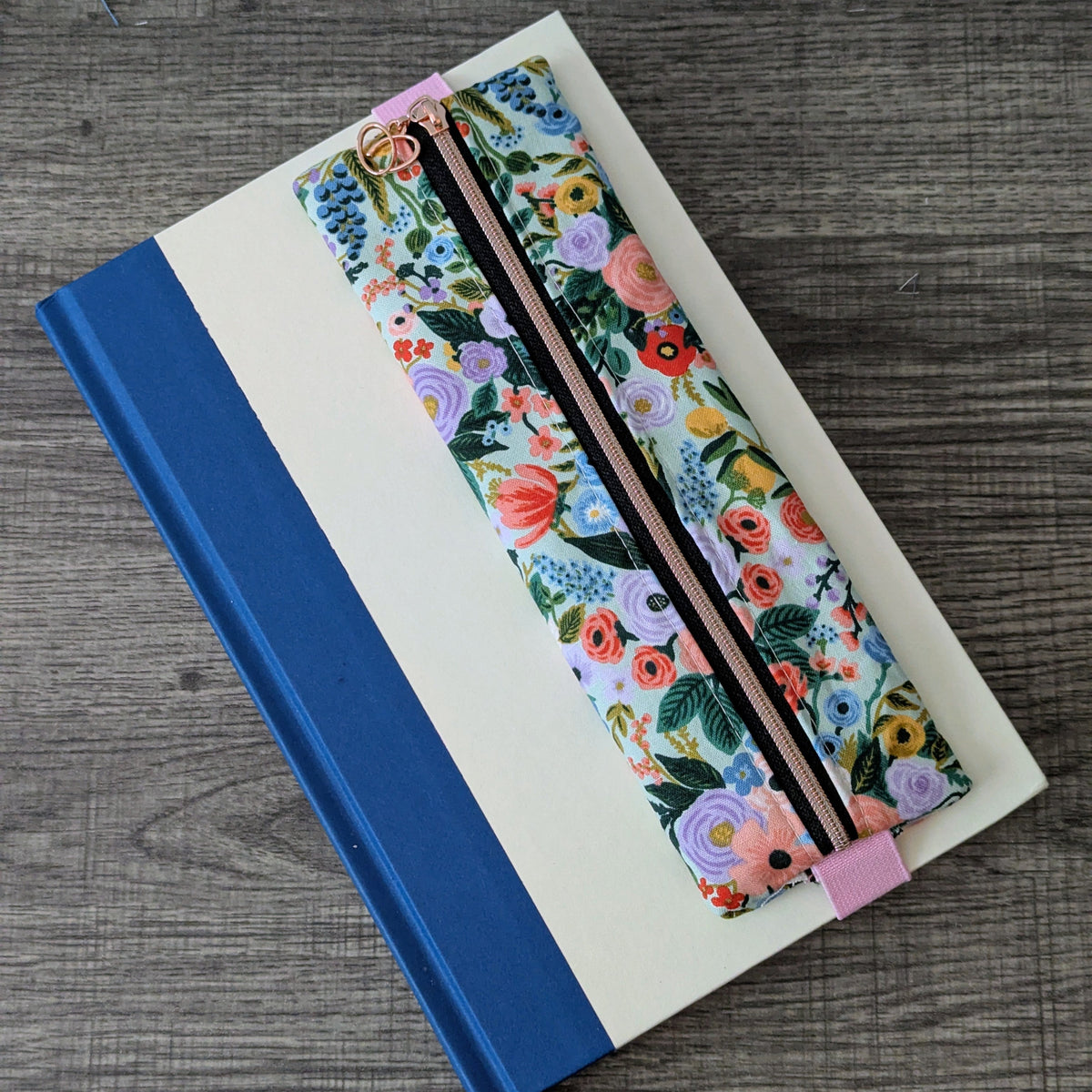 A floral-patterned pencil case with a black zipper and pink elastic band, placed on top of a cream-colored notebook with a blue spine, set on a gray wood-textured surface.