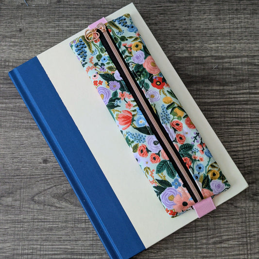 A floral-patterned pencil case with a black zipper and pink elastic band, placed on top of a cream-colored notebook with a blue spine, set on a gray wood-textured surface.