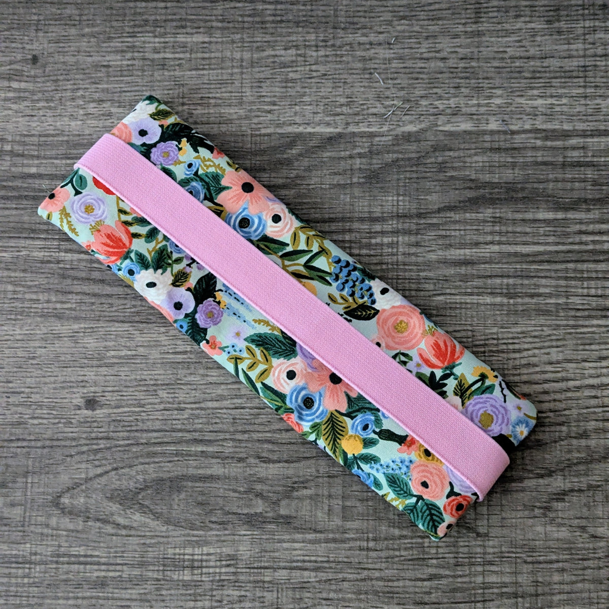 A fabric zippered pouch with a colorful floral print and a wide pink elastic band, shown on a gray wood-textured background.