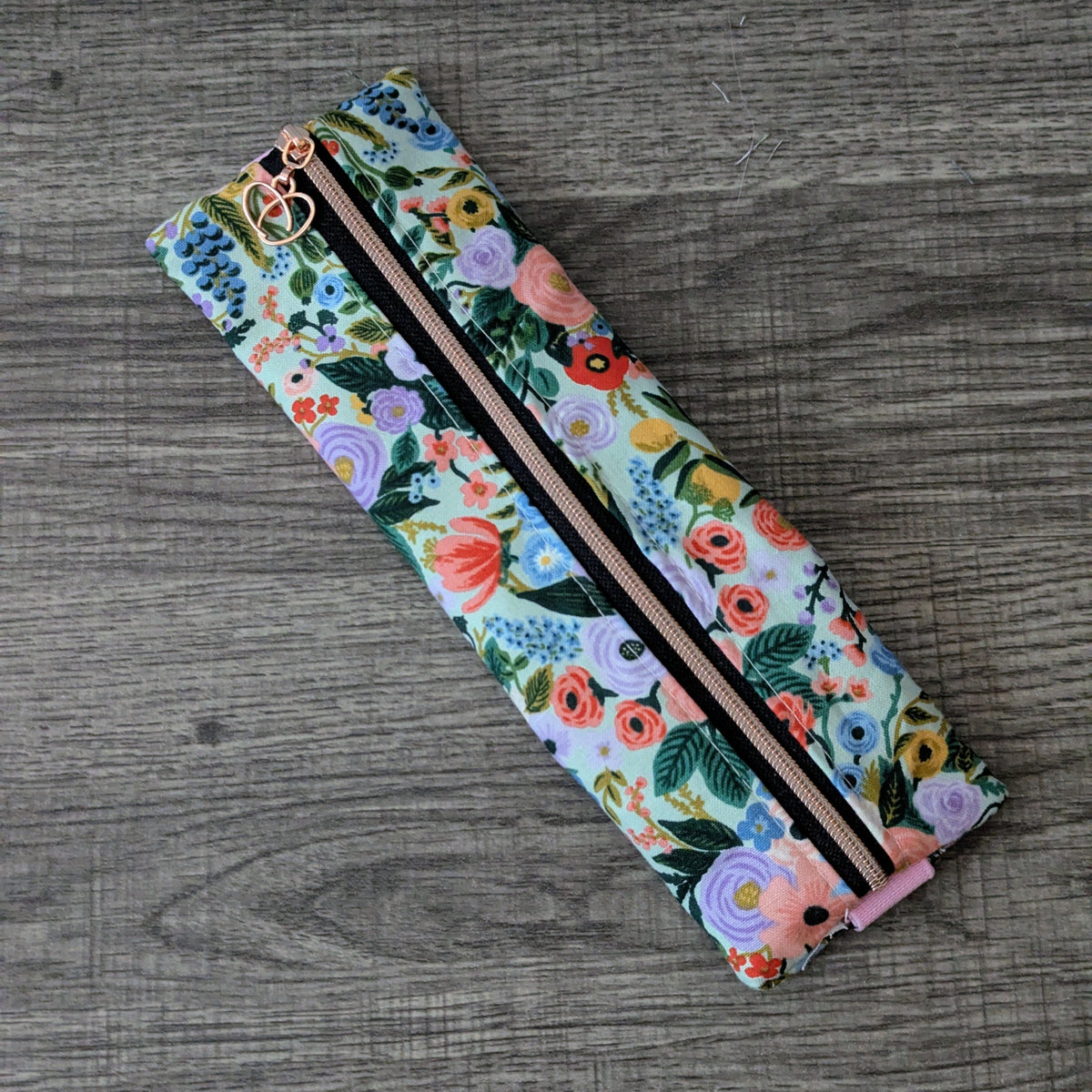 A long, narrow fabric pouch or case with a colorful floral print, laid diagonally on a gray wood-textured surface. The print features pink, purple, yellow, and green flowers on a light blue background, with a black zipper running down the center and a pink elastic band.