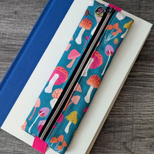 A zippered fabric pencil case with a colorful mushroom print on a teal background, resting diagonally on top of a white notebook with a blue spine on a gray wood-textured surface.