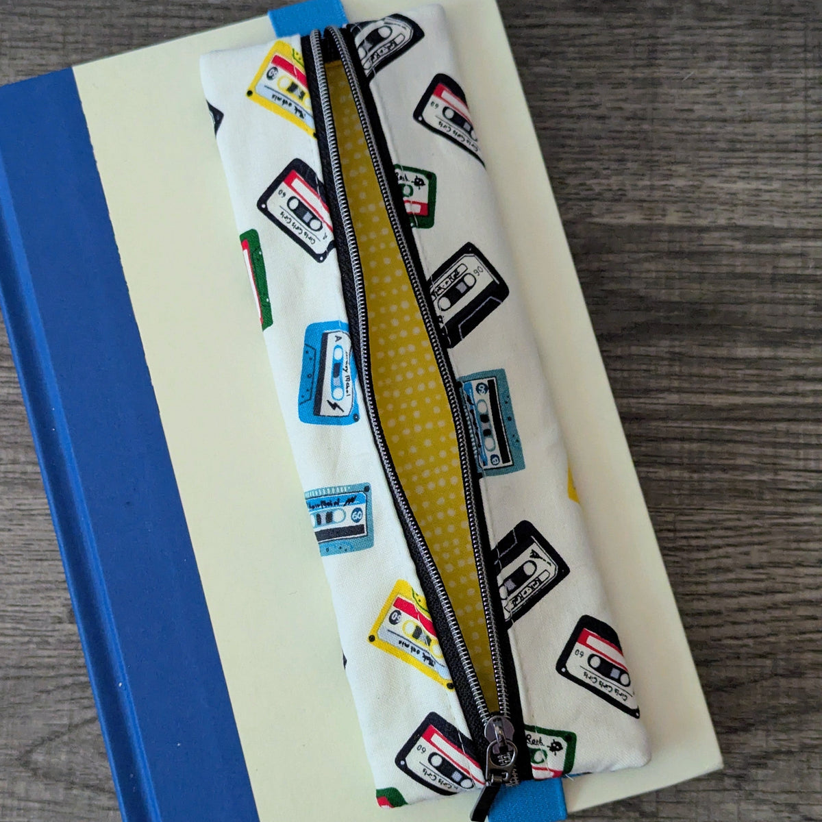 A white fabric pencil case with a black zipper and a colorful cassette tape pattern, resting diagonally on top of a cream and blue notebook over a gray wood-grain surface.