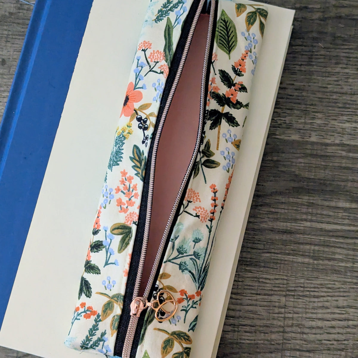 A floral zippered pencil case with a rose gold zipper and charm, resting on top of a notebook on a gray wood-grain surface.