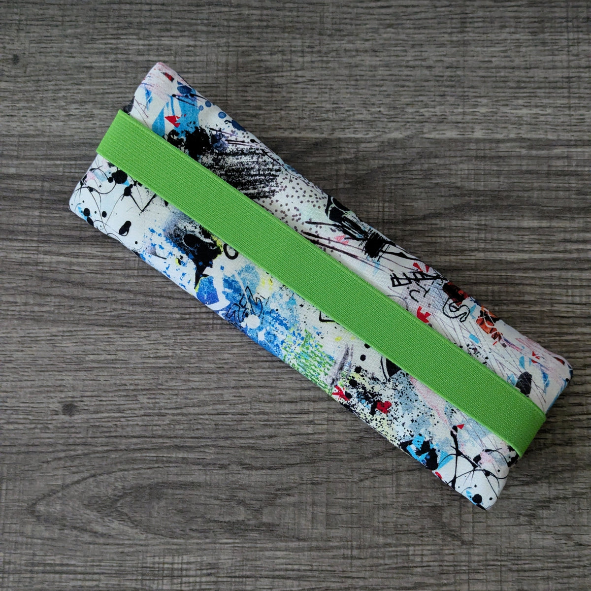 A fabric zippered pouch with a colorful abstract paint splatter pattern and a bright green elastic band, shown on a gray wood-textured background.