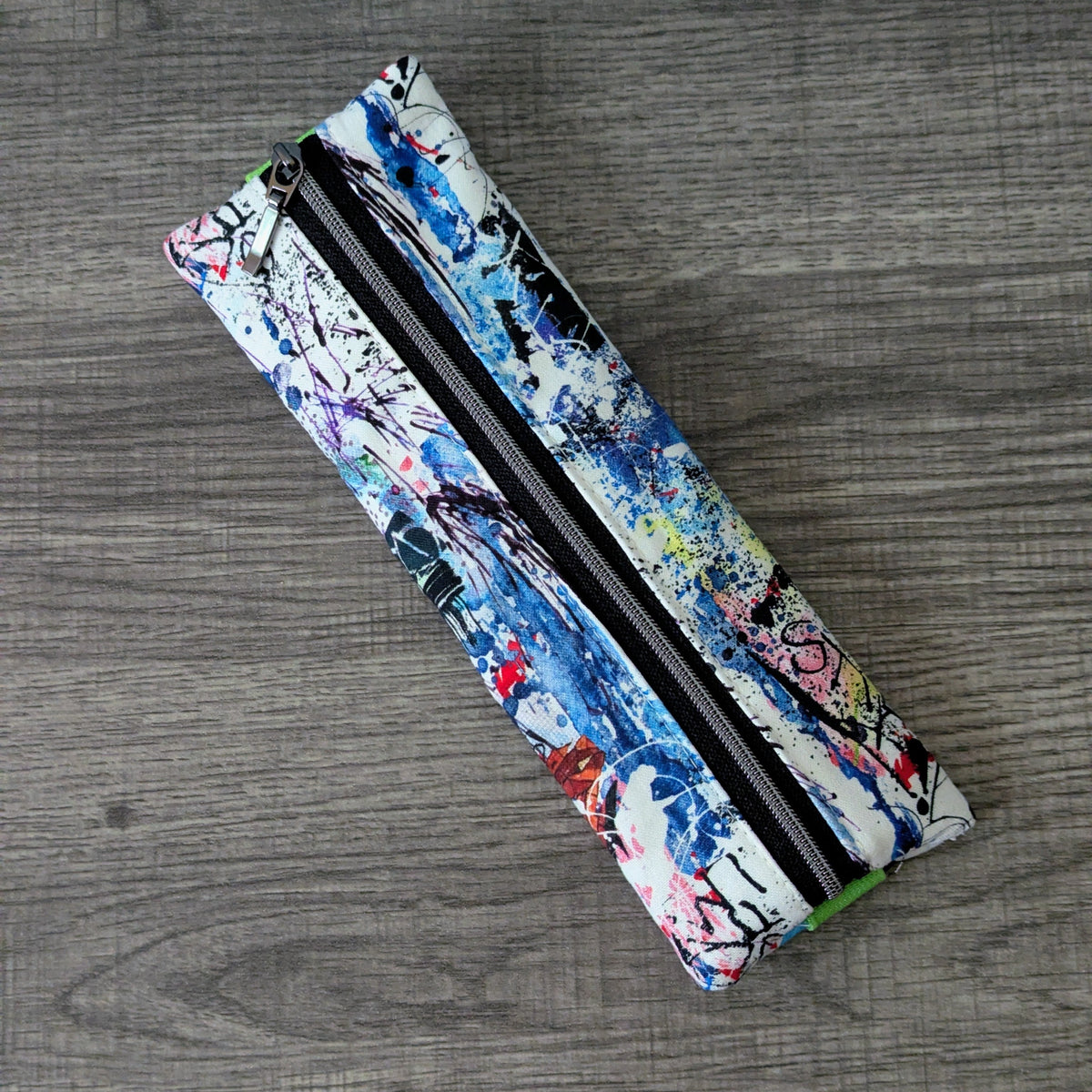 A rectangular zippered pouch with a colorful abstract paint-splatter pattern, placed diagonally on a gray wood-textured surface.