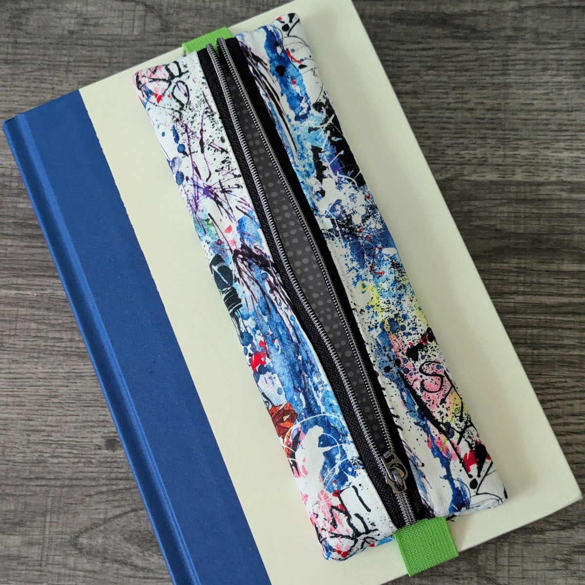 A zippered pencil case with a colorful abstract paint splatter pattern, resting diagonally on top of a cream-colored notebook with a blue spine, set on a gray wood-textured surface.
