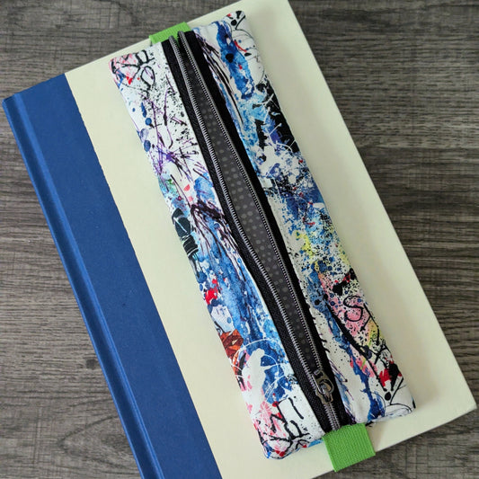 A zippered pencil case with a colorful abstract paint splatter pattern, resting diagonally on top of a cream-colored notebook with a blue spine, set on a gray wood-textured surface.