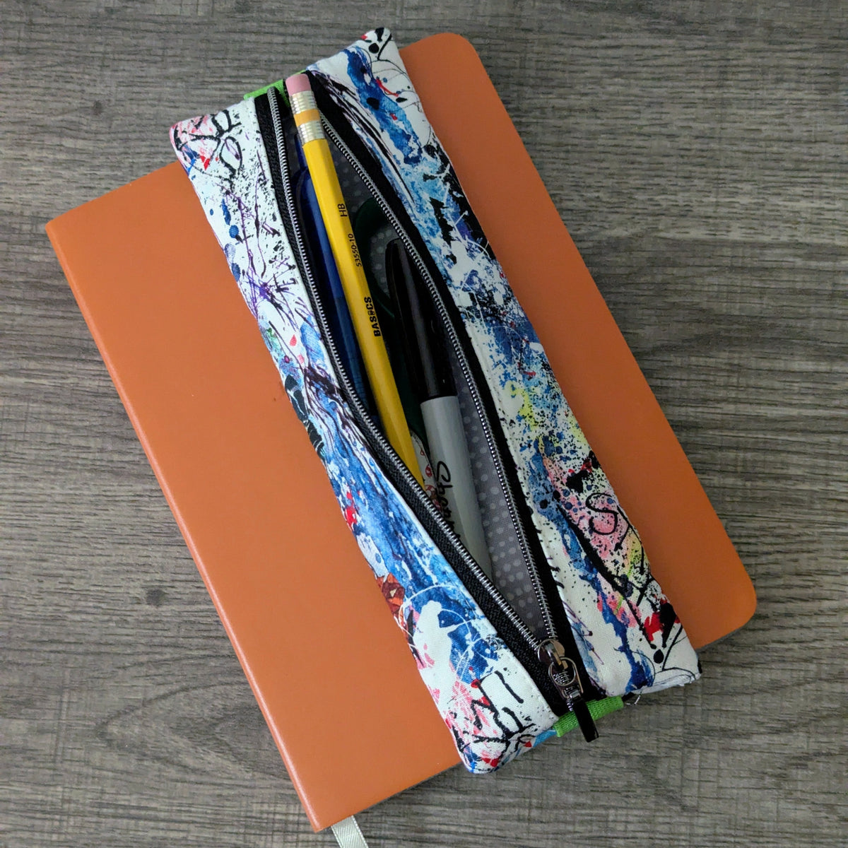 A zippered pencil pouch with a colorful graffiti-style paint splatter pattern, shown open on top of an orange notebook over a gray wood-grain surface.