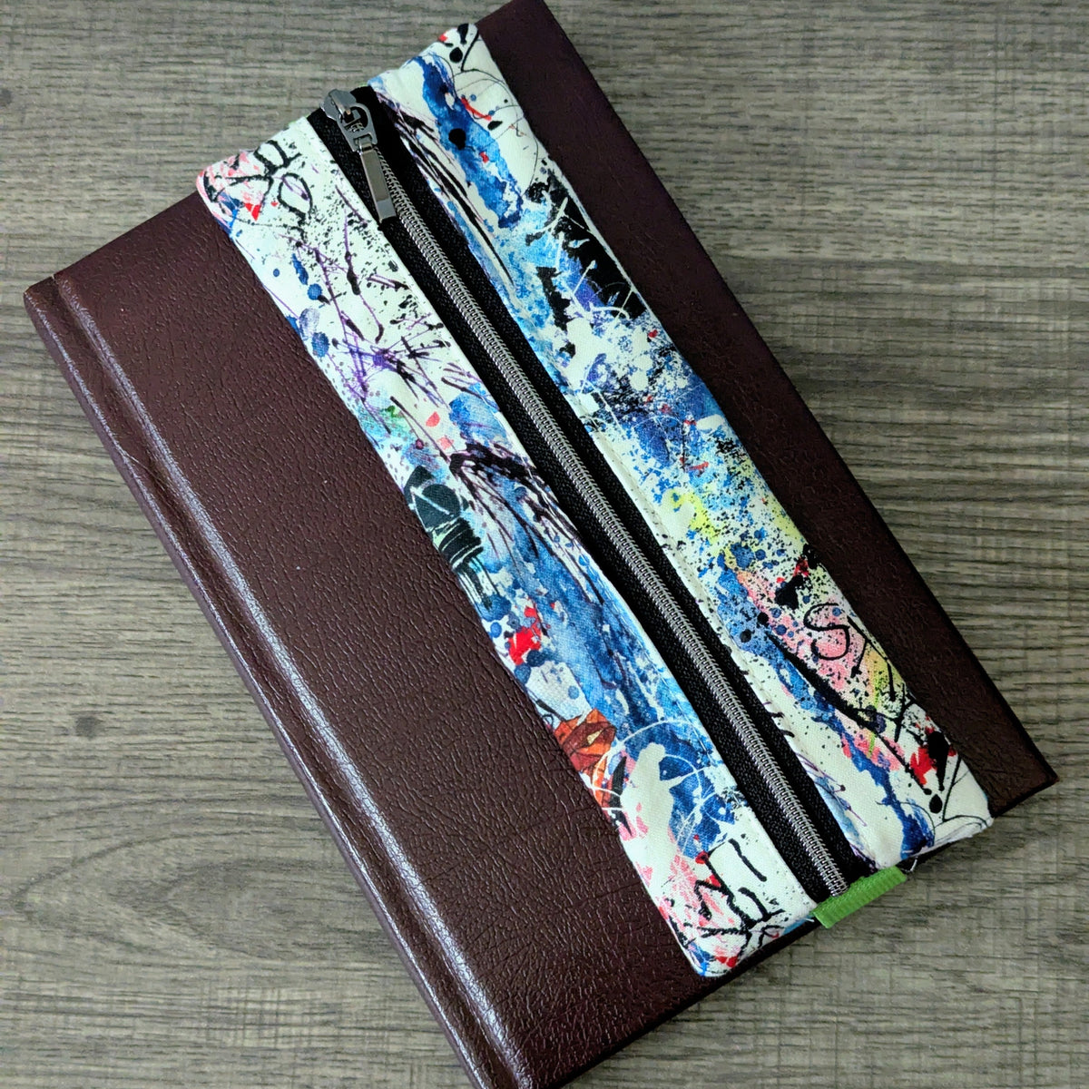 A dark brown leather-bound notebook or journal with a colorful abstract graffiti-patterned elastic closure pencil case, placed on a gray wood-textured surface.