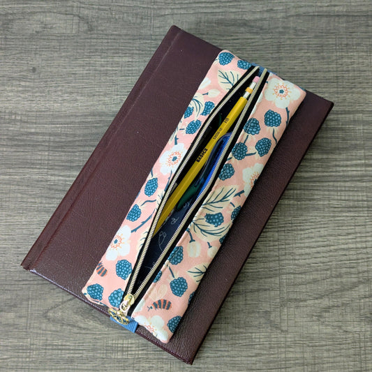 A pink zippered pencil case with a blue berry and leaf pattern, shown open with pens inside on top of a dark brown leather notebook, set on a gray wood-grain surface.