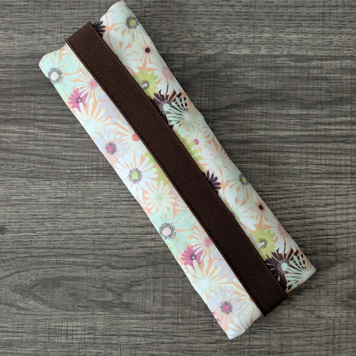 A fabric zippered pouch with a brown elastic band, featuring a light pastel floral and butterfly pattern, laid diagonally on a gray wood-textured background.