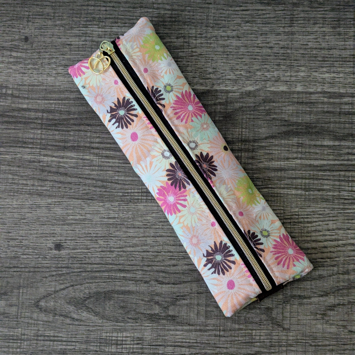 A slim zippered pouch with a colorful floral print, laid diagonally on a gray wood-textured surface.