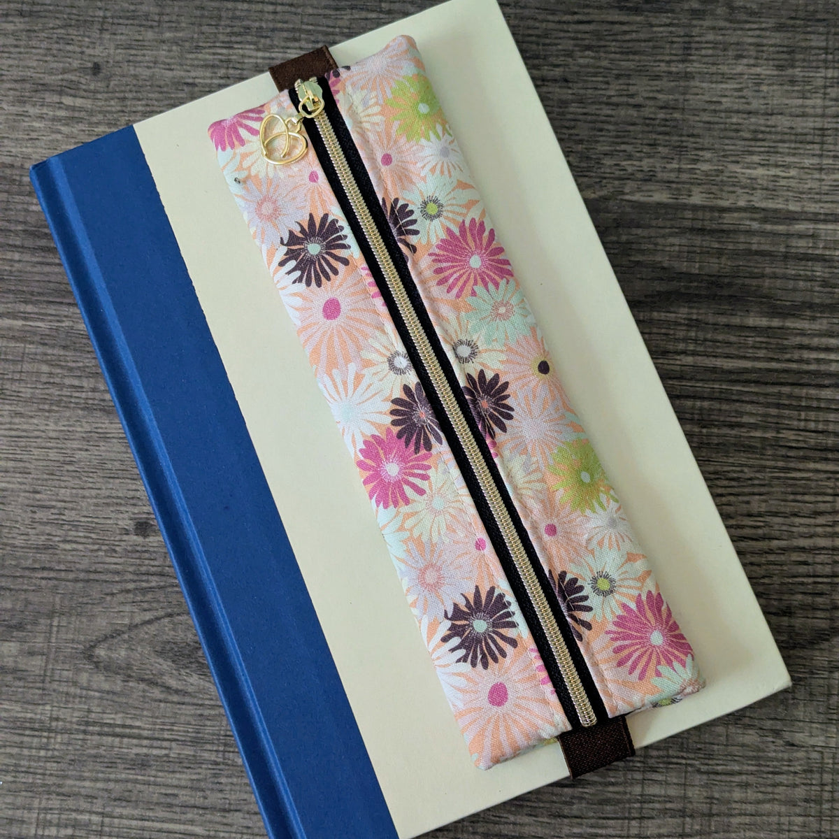 A floral zippered pencil case with pink, purple, and green flower patterns, resting on top of a cream-colored notebook with a blue spine, placed on a gray wood-textured surface.