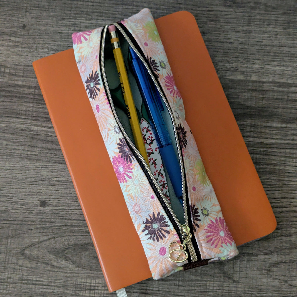 A floral zippered pencil pouch containing pens and a pencil, resting diagonally on top of an orange notebook over a gray wood-grain surface.
