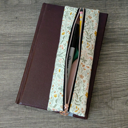 A rectangular zip pouch with a floral print lining and a dark brown faux-leather exterior, shown open on a gray wood-grain tabletop. Pens and pencils are visible inside the pouch.